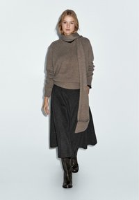 Kvinde går iført brun sweater med matchende tørklæde, mørk tekstureret midi-nederdel og sorte knæhøje støvler mod en ensfarvet baggrund.