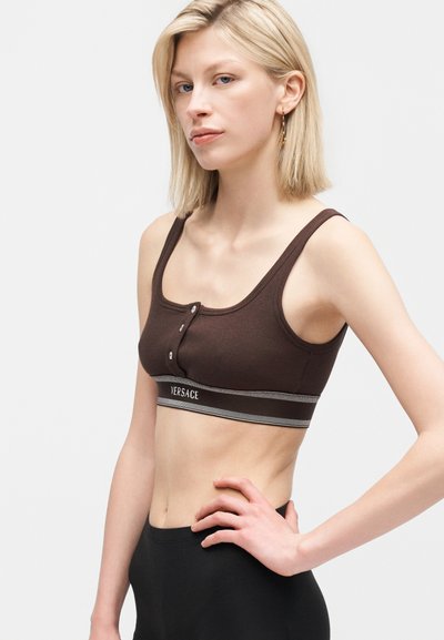 Ξανθιά γυναίκα που φοράει ένα καφέ crop top Versace με κουμπιά, όρθια με το χέρι στη μέση μπροστά σε απλό ανοιχτό φόντο.