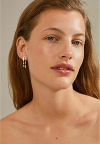 Orecchini a goccia in argento con un design curvo, caratterizzati da tre catene a cascata e accenti circolari. Modella con capelli di lunghezza media e trucco naturale.