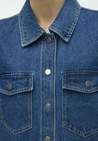 Denimjakke med kragehals, to frontlommer med klaffer, sølvfargede trykknapper og en mellomblå vask med kontraststing.