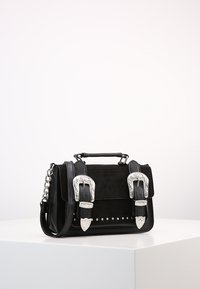 Borsa in pelle nera e suede con fibbie incise in argento, dettagli con borchie, manico superiore e tracolla a catena su superficie bianca.