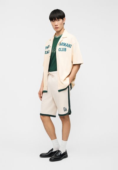 Μπεζ κοντομάνικο πουκάμισο με πράσινο κείμενο, συνδυασμένο με σκούρο πράσινο μπλουζάκι και μπεζ σορτς με πράσινη επένδυση και λογότυπο. Μαύρα παπούτσια και λευκές κάλτσες.