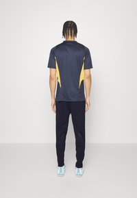 Camiseta atlética de color marino con acentos laterales amarillos, combinada con pantalones oscuros. La camiseta tiene cuello redondo y mangas cortas; los pantalones cuentan con paneles texturizados.