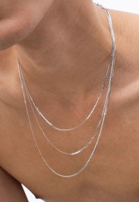 Silberne Kettenhalskette mit drei geschichteten Strängen; dünne, flache Glieder sorgen für eine glatte Textur. Dekolleté sichtbar, heller Hautton.