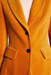 Corduroy blazer i lys orange med klippede revers, to forlommer og sorte knapper, der fremhæver en ribbet tekstur.