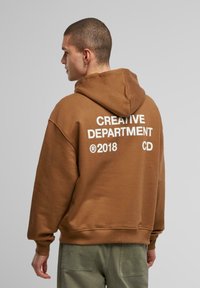 Brun hættetrøje lavet af blødt stof, med hvid tekst "CREATIVE DEPARTMENT" og "®2018 CD" på ryggen, samt ribbede manchetter.