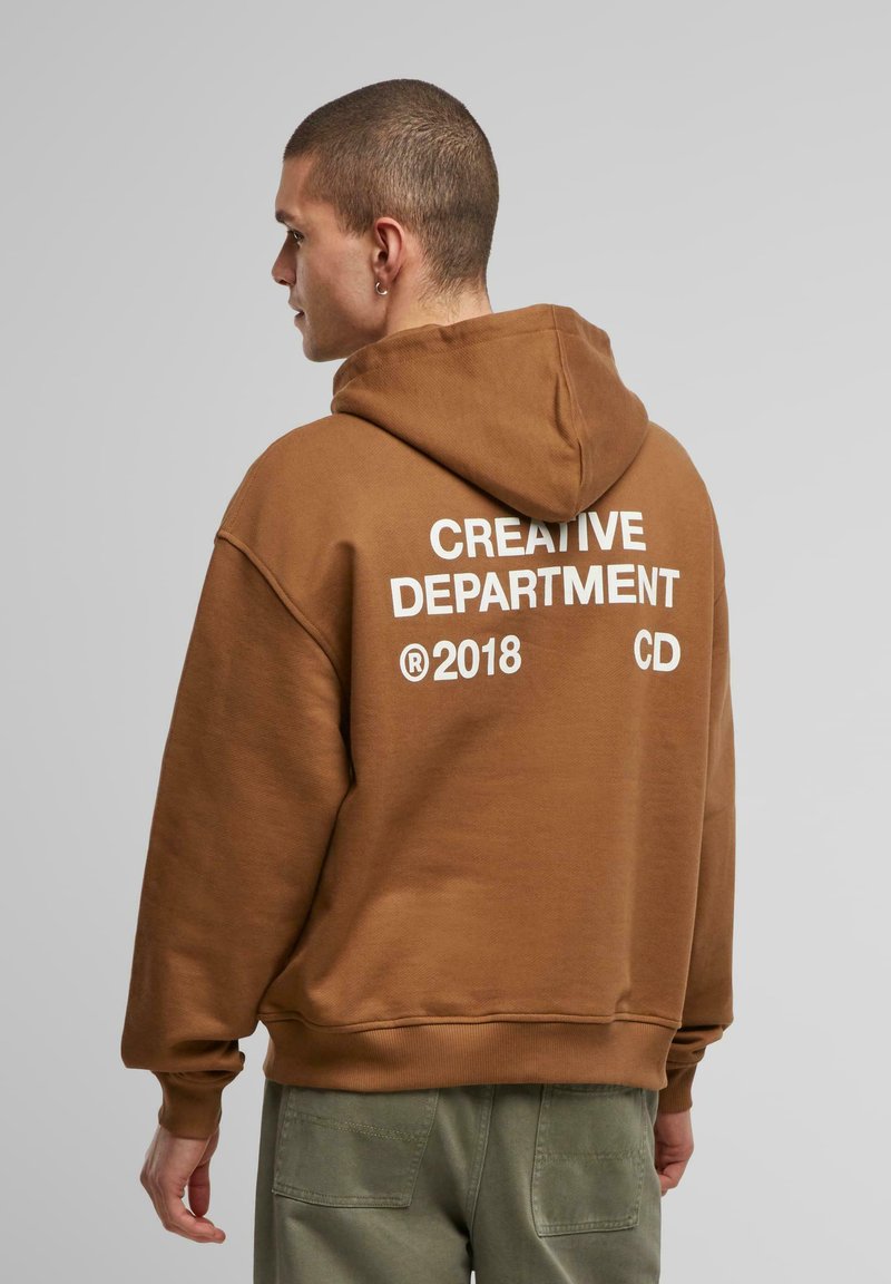Brun hættetrøje lavet af blødt stof, med hvid tekst "CREATIVE DEPARTMENT" og "®2018 CD" på ryggen, samt ribbede manchetter.