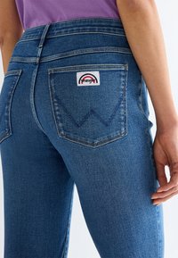 Prim-plan cu o persoană care poartă blugi Wrangler albaștri și un tricou mov, arătând buzunarul din spate cu patch-ul logo Wrangler.