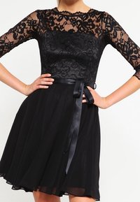 Svart klänning med en spetsöverdel, rund halsringning, trekvartslånga ärmar och en flödande chiffongsjurk, avslutad med ett satinbälte.