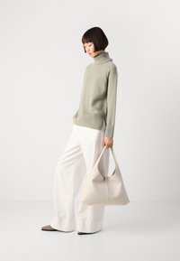 Woman wearing a light gray turtleneck sweater and white wide-leg pants, holding a large cream-colored handbag, standing against a plain backdrop.