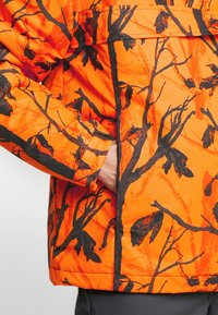 Giacca arancione brillante con un motivo di ramificazioni nere. Il materiale appare liscio, con una tasca con zip e un cordino regolabile all'orlo.