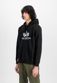 Felpa nera con logo bianco che riporta la scritta "Alpha Industries". Presenta una tasca frontale, cappuccio con stringhe e polsini a costine. Il modello indossa pantaloni cargo verdi oliva.