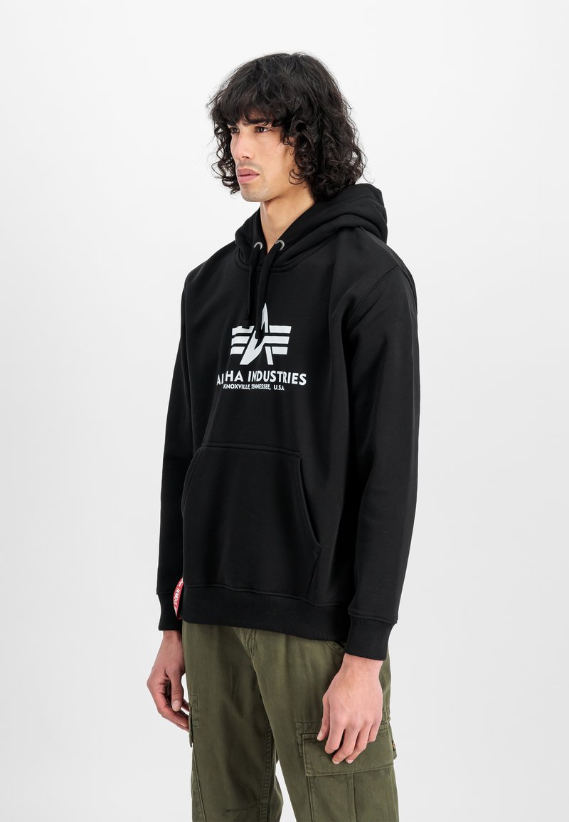 Felpa nera con logo bianco che riporta la scritta "Alpha Industries". Presenta una tasca frontale, cappuccio con stringhe e polsini a costine. Il modello indossa pantaloni cargo verdi oliva.