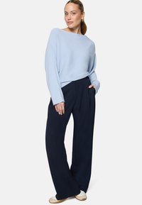 Femme portant un pull oversize bleu clair, un pantalon large bleu marine, des baskets blanches et des boucles d'oreilles créoles dorées, debout avec une main dans la poche.