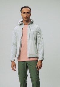 Lichtgrijze waterdichte jas met een capuchon, voorzien van een rits aan de voorkant, gedragen over een lichtroze shirt, gecombineerd met groene cargobroek.