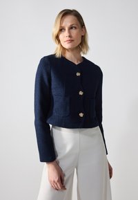 Veste cropped navy en tissu texturé, avec des accents de boutons en forme de fleurs dorées et deux poches avant. Portée sur un pantalon blanc.
