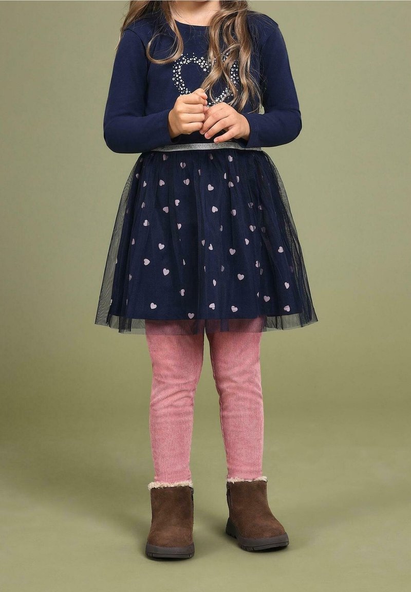 Enfant portant une robe manches longues bleu marine avec des motifs en forme de cœur, des leggings roses et des bottes marron, debout devant un fond olive uni.