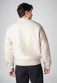 Cream-colored fluffy bomber jacket with ribbed cuffs and hem, featuring a relaxed fit and minimalistic design against a plain background.