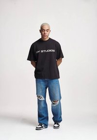 T-shirt oversized preta com "2Y STUDIOS" impresso em branco, combinado com jeans claros desgastados apresentando rasgos nos joelhos e tênis pretos.