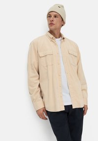 Licht beige overhemd met knopen, voorzien van twee borstzakken en een ontspannen pasvorm, gelaagd over een wit shirt, gecombineerd met donkere broek en een crèmekleurige beanie.