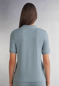 Světle šedomodré polo tričko s límečkem, krátkými rukávy a bílými lemovkami podél límce a manžet. Hladká textura.