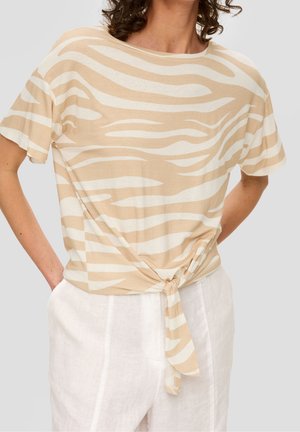 Kobieta w beżowo-białym, krótkim topie w paski zebry, zawiązanym w talii, w połączeniu z białymi lnianymi spodniami na jasnym tle.