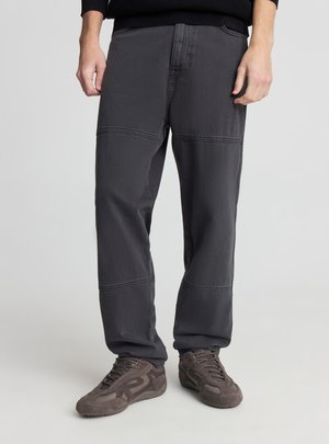 Mężczyzna ubrany w ciemnoszare spodnie z przeszyciami i brązowe zamszowe trampki, stojący na jasnym, gładkim tle.