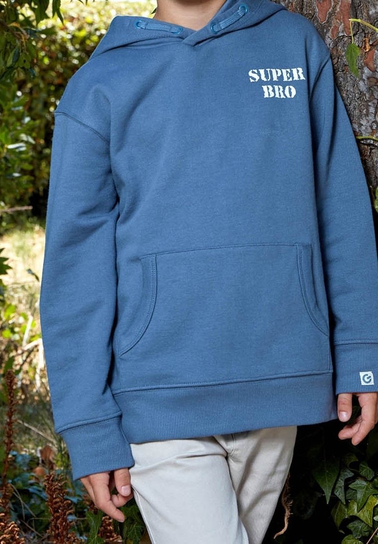 Enfant portant un sweat à capuche bleu avec le texte "SUPER BRO", debout à l'extérieur près d'un arbre avec un feuillage vert en arrière-plan.