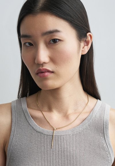 Χρυσό κολιέ με σχέδιο σε σχήμα Y που κρέμεται από μια λεπτή αλυσίδα, φορεμένο πάνω από μια γκρι ριμπ μπλούζα.