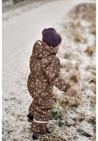 Combineu de zăpadă maro cu model floral, cu glugă și accente albe, asortat cu o căciulă mov și mănuși bej, purtat de un copil într-un decor înzăpezit.
