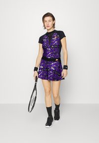 Lila und schwarze Tennisanzug mit einem taillierten Oberteil mit kunstvollen Mustern und einem ausgestellten Rock, ergänzt durch einen Tennisschläger.
