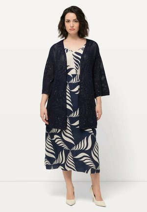 Navy open-knit kimono cardigan over a navy and cream leaf-patterned dress, with short sleeves and a relaxed fit. Cream-colored heels.