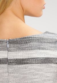 Texturované šaty v šedých, bílých a černých pruzích. Mají kulatý výstřih a skrytý zip na zadní straně pro zapínání.