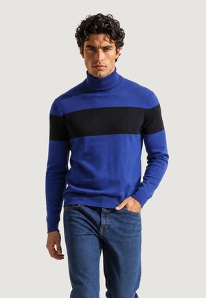 Uomo con un maglione dolcevita blu con una larga striscia orizzontale nera e jeans blu, in piedi con una mano in tasca.