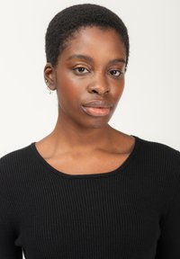 Schwarzer Rippstrickpullover mit weitem Ausschnitt. Das Model hat kurzes, natürliches Haar und trägt einen kleinen silbernen Ohrring. Der Hautton ist warm.