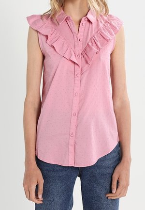 Ärmellose rosa Bluse mit Knöpfen vorne und gerüschten V-förmigen Details, getragen zu blauen Jeans.