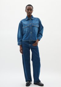 Giacca di jeans di blu medio con tasche sul petto e chiusura con bottoni, abbinata a pantaloni in denim a gamba ampia, sempre di blu medio. Scarpe nere.