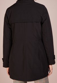 Svart vattentät kappa med ryggpanel, justerbara ärmslut och en slät yta. Har rak skärning och dolda ficköppningar.