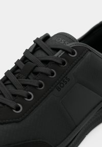 Κοντινή λήψη ενός μαύρου αθλητικού παπουτσιού με υφή ύφασμα, κορδόνια σε ίδιο χρώμα και τη σήμανση "BOSS" στη γλώσσα και στην πλευρική επιφάνεια.