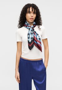 Μαντήλι από μετάξι με γεωμετρικό μοτίβο σε μπλε, μαύρο και μπορντό. Δεμένο γύρω από τον λαιμό με κόμπο, που συμπληρώνει ένα ανοιχτόχρωμο τοπ.