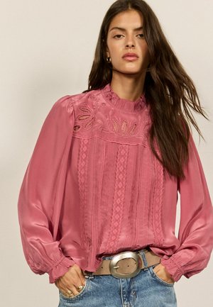 Kvinde med langt mørkt hår iført en pink blondebluse med puffede ærmer, stoppet ind i blå jeans med en stor sølvspænde.