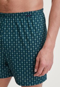 Boxers pour hommes en tissu bleu canard avec un motif géométrique aux accents vert clair et bleu. Ceinture élastique avec étiquette de logo.