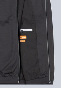 Schwarze Jacke mit tailliertem Design, ausgestattet mit einer Reißverschlusstasche, reflektierenden Akzenten und einem orangefarbenen Markenlabel. Glatte, gewebte Textur.