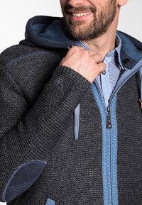 Hooded jack in donkergrijze stof met blauwe accenten. Voorzien van een gestructureerde weving, ritssluiting en elleboogpatches. Zachte binnenvoering zichtbaar.