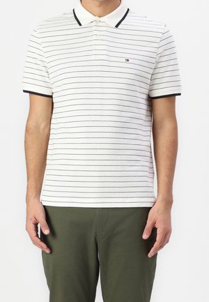 Homme portant un polo blanc à manches courtes avec de fines rayures horizontales noires et un pantalon vert foncé.