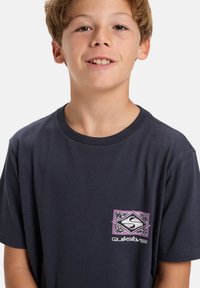 Dark navy cotton t-shirt featuring a colorful Quiksilver logo patch on the left chest, round neckline, and short sleeves.