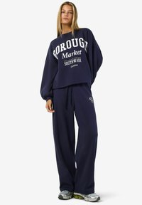 Tumši zilas krāsas pārāk liela izmēra jakas ar baltu grafisku tekstu un saderīgām platajām sporta biksēm, izgatavotas no mīksta auduma, kombinētas ar sudraba sporta apaviem.