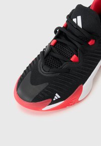 Baskets de sport noires avec des accents rouges, tige en mesh, surface texturée, logo proéminent et détails blancs. Design élégant avec une semelle en caoutchouc.