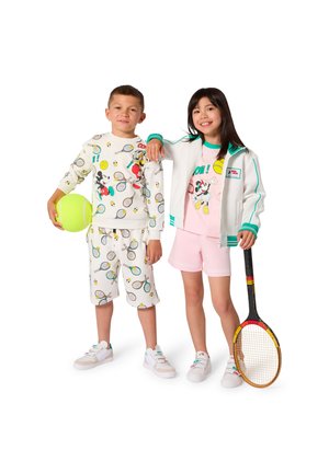 Due bambini con abiti a tema tennis, uno tiene una grande pallina da tennis, l'altro una racchetta da tennis, sorridendo su sfondo bianco.