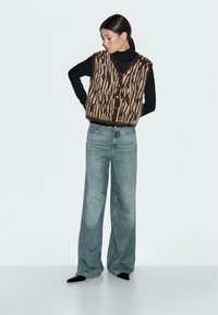 Brun och beige mönstrad stickad väst med V-ringning och knappar, bärs över en svart långärmad skjorta och matchas med ljusblå vida jeans.